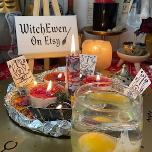 Puede incluir: Un vaso transparente con un huevo crudo en agua se encuentra sobre una bandeja circular dorada con un pequeño caballete de madera que muestra un letrero que dice WitchEwen On Etsy. Varias velas encendidas de colores y pequeñas tarjetas de papel con dibujos en tinta negra están dispuestas en la bandeja, que está forrada con papel de aluminio y pétalos de flores secas esparcidos.