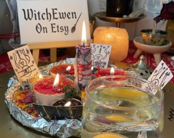 May include: A clear glass containing a raw egg in water sits on a gold-toned circular tray with a small wooden easel displaying a sign that reads WitchEwen On Etsy. Several lit candles in various colors and small paper cards with black ink drawings are arranged on the tray, which is lined with aluminum foil and scattered dried flower petals.