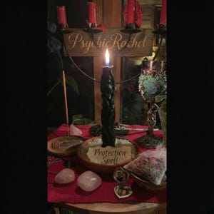 May include: A dimly lit altar with a burning black candle as the focal point. The scene includes a wooden sign that reads "Psychic Rachel", a small bowl with the words "Protection Spell", and other ritualistic items, creating a mystical atmosphere.