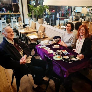 Puede incluir: Escena de caf&eacute; con personas reunidas alrededor de una mesa cubierta con un pa&ntilde;o morado. Se ven cartas del tarot, tazas y un platillo. El escenario incluye mesas de madera, sillas y plantas decorativas.