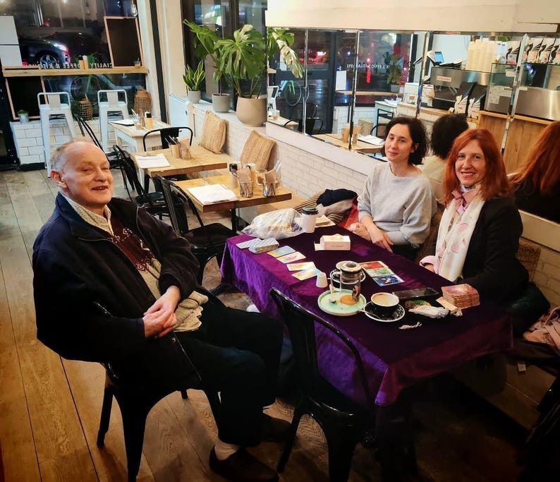 Puede incluir: Escena de caf&eacute; con personas reunidas alrededor de una mesa cubierta con un pa&ntilde;o morado. Se ven cartas del tarot, tazas y un platillo. El escenario incluye mesas de madera, sillas y plantas decorativas.