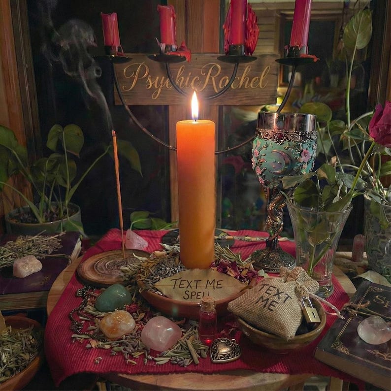 Puede incluir: Una vela naranja encendida es el punto focal, rodeada de cristales, hierbas y una copa decorativa. Una peque&ntilde;a bolsa de yute con "TEXT ME" y un letrero de madera que dice "TEXT ME SPELL" tambi&eacute;n est&aacute;n presentes. La escena sugiere un tema espiritual o m&iacute;stico.