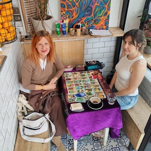 Puede incluir: Dos mujeres sentadas en una mesa cubierta con un pa&ntilde;o morado, mostrando cartas del tarot. Una taza de caf&eacute; y un vaso de agua est&aacute;n sobre la mesa. El escenario es un caf&eacute;, con naranjas en una cesta y obras de arte coloridas.