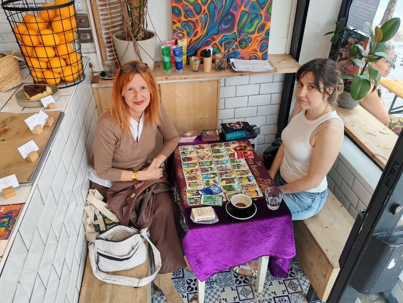 Puede incluir: Dos mujeres sentadas en una mesa cubierta con un pa&ntilde;o morado, mostrando cartas del tarot. Una taza de caf&eacute; y un vaso de agua est&aacute;n sobre la mesa. El escenario es un caf&eacute;, con naranjas en una cesta y obras de arte coloridas.