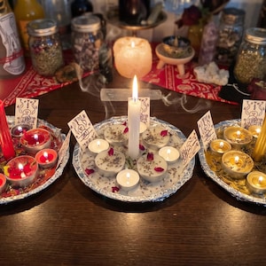 Peut inclure: Trois plateaux ronds en papier aluminium contenant des bougies allumées et des herbes séchées. Le plateau de gauche a une bougie effilée rouge, celui du centre une blanche et celui de droite une dorée, avec des bougies chauffe-plat et des cartes.
