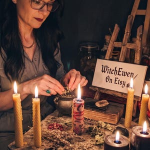 May include: A collection of ritual items including a small metal cauldron, several beeswax candles, two black pillar candles with etched runes, dried herbs, and a sign on a wooden easel that reads WitchEwen/On Etsy, arranged on a stone surface.