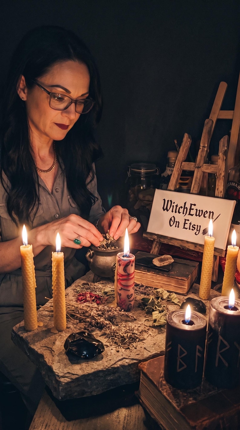 May include: A collection of ritual items including a small metal cauldron, several beeswax candles, two black pillar candles with etched runes, dried herbs, and a sign on a wooden easel that reads WitchEwen/On Etsy, arranged on a stone surface.