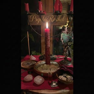 May include: A central lit red candle is surrounded by other candles, crystals, and a chalice. The text "Psychic Rachel" is visible above the main candle. The words "Think of Me" are written on a wooden bowl. The scene is set on a red cloth.