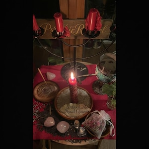 Può includere: Un altare illuminato da candele con una candela rossa accesa, cristalli di quarzo rosa e un piccolo sacchetto di fiori secchi. Un cartello di legno con le lettere "Psic R" è visibile sullo sfondo. Il testo "Unblock Me Road Opener" è su un piatto di legno.
