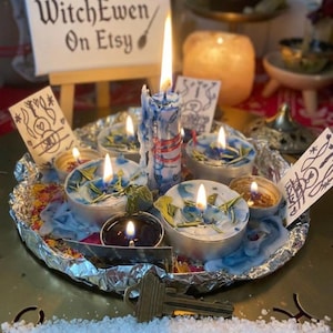 Puede incluir: Una colección de velas de té encendidas y una vela de pilar más alta con gotas de cera azul, dispuestas en una bandeja forrada con papel de aluminio y hierbas secas. Una llave de color latón descansa al frente, con sal blanca y tarjetas de papel.