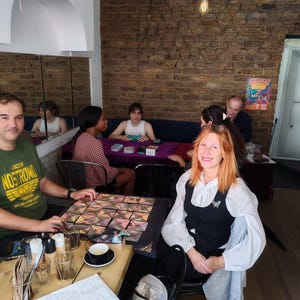 Puede incluir: Un grupo de personas reunidas alrededor de mesas en un caf&eacute;. Algunos juegan un juego de mesa con fichas geom&eacute;tricas de colores. Otros est&aacute;n sentados, algunos con cartas. El caf&eacute; tiene una pared de ladrillo y un espejo.