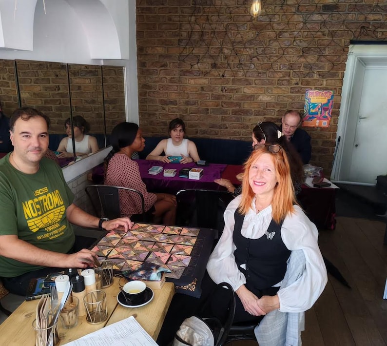 Puede incluir: Un grupo de personas reunidas alrededor de mesas en un caf&eacute;. Algunos juegan un juego de mesa con fichas geom&eacute;tricas de colores. Otros est&aacute;n sentados, algunos con cartas. El caf&eacute; tiene una pared de ladrillo y un espejo.