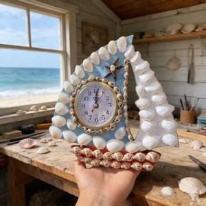 Könnte beinhalten: Eine handgefertigte Segelbootuhr, verziert mit Muscheln. Das Zifferblatt ist rund mit römischen Ziffern und einer goldfarbenen Lünette. Das Segel ist hellblau und der Bootsrumpf rot. Ein Seestern schmückt die Spitze des Segels.