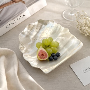 May include: A pearlescent, shell-shaped serving dish holds green grapes, a sliced fig, and blueberries. The dish has a wavy edge and sits on a neutral surface. A magazine with the word "KINFOLK" is partially visible.