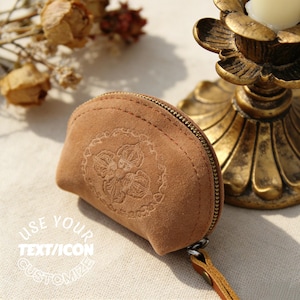 May include: A small, brown suede coin purse with a floral design embossed on the front. The purse has a curved shape, a metal zipper, and a leather pull tab. The text "USE YOUR TEXT/ICON CUSTOMIZE" is visible.