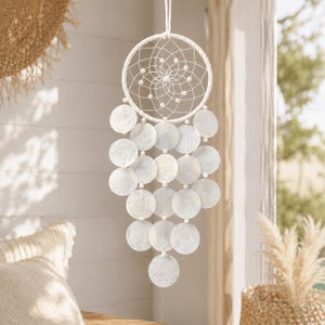 May include: A white dream catcher with a circular woven frame and a webbed center, adorned with small white beads. Below the frame, multiple round, iridescent shell discs hang in rows, creating a decorative and airy design.
