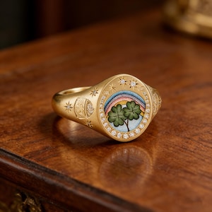 Puede incluir: Un anillo de oro con un diseño detallado. El anillo presenta un óvalo central con un arcoíris y dos tréboles de cuatro hojas. El óvalo está rodeado de pequeños diamantes. El anillo también tiene detalles de estrellas y luna.