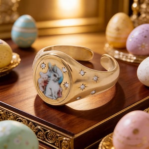 Puede incluir: Un brazalete dorado con un diseño ovalado central que presenta una imagen pintada de un conejo y una figura. La pulsera está adornada con detalles en forma de estrella y pequeños diamantes. El fondo incluye huevos de Pascua decorados.