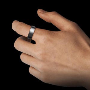 May include: A black and silver ring with a flat band is worn on a finger. The ring has a polished silver top and a matte black band. The background is solid black.