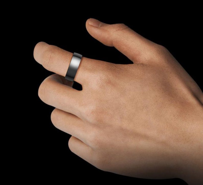 May include: A black and silver ring with a flat band is worn on a finger. The ring has a polished silver top and a matte black band. The background is solid black.