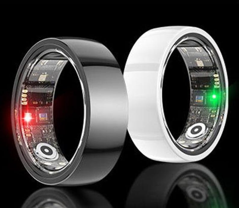 May include: Two smart rings, one black and one white, are displayed against a black background. The rings have a sleek, modern design with a visible circuit board and embedded lights. The black ring has a red light, and the white ring has a green light.