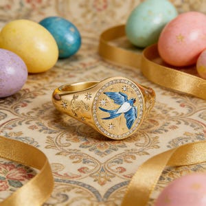 May include: A gold ring with a blue and white bird design, surrounded by small diamonds. The ring is on a patterned surface with colorful Easter eggs and gold ribbon.