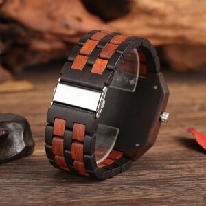 Puede incluir: Un reloj de pulsera de madera con una esfera rectangular y una correa hecha de segmentos de madera marr&oacute;n oscuro y claro alternados. El reloj presenta un cierre plateado y una esfera oscura. El reloj se muestra sobre una superficie de madera.