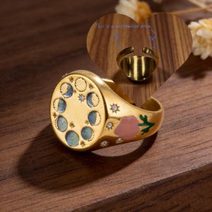 Può includere: Anello dorato con un design dettagliato di fasi lunari e stelle. L'anello presenta un accento floreale con smalto rosa e verde. Il testo nell'immagine dice: "This is a customized area."