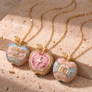 Könnte beinhalten: Drei goldene Halsketten mit apfelförmigen Anhängern. Jeder Anhänger ist rosa mit einem anderen Design: ein Karussell, eine Ballerina und ein Karussell. Die Anhänger haben goldene Blattdetails und liegen auf einer strukturierten Oberfläche.