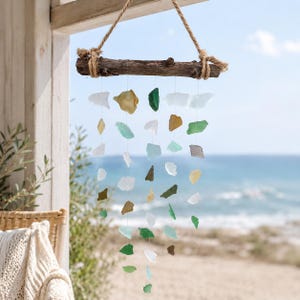 Può includere: Un carillon decorativo realizzato con un ramo di legno stagionato e fili di vetro marino. I pezzi di vetro marino sono di vari colori, tra cui bianco, verde, marrone e ambra, e sono sospesi da fili trasparenti. Il carillon è appeso a una corda naturale.