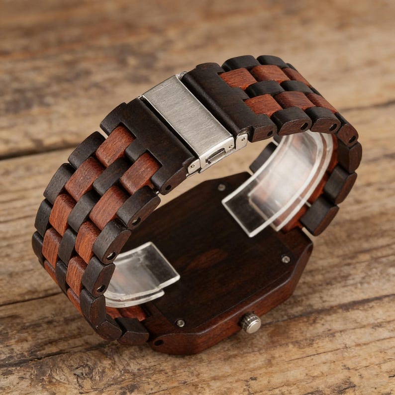 Puede incluir: Un reloj de pulsera de madera con una esfera rectangular marr&oacute;n oscuro y una correa hecha de eslabones de madera marr&oacute;n oscuro y marr&oacute;n rojizo alternados. El reloj tiene un cierre plateado y una pieza de pl&aacute;stico transparente.