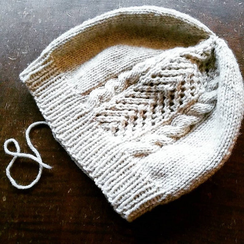 Puede incluir: Un gorro de punto blanco con un patr&oacute;n de trenza. El gorro est&aacute; sobre una superficie de madera.