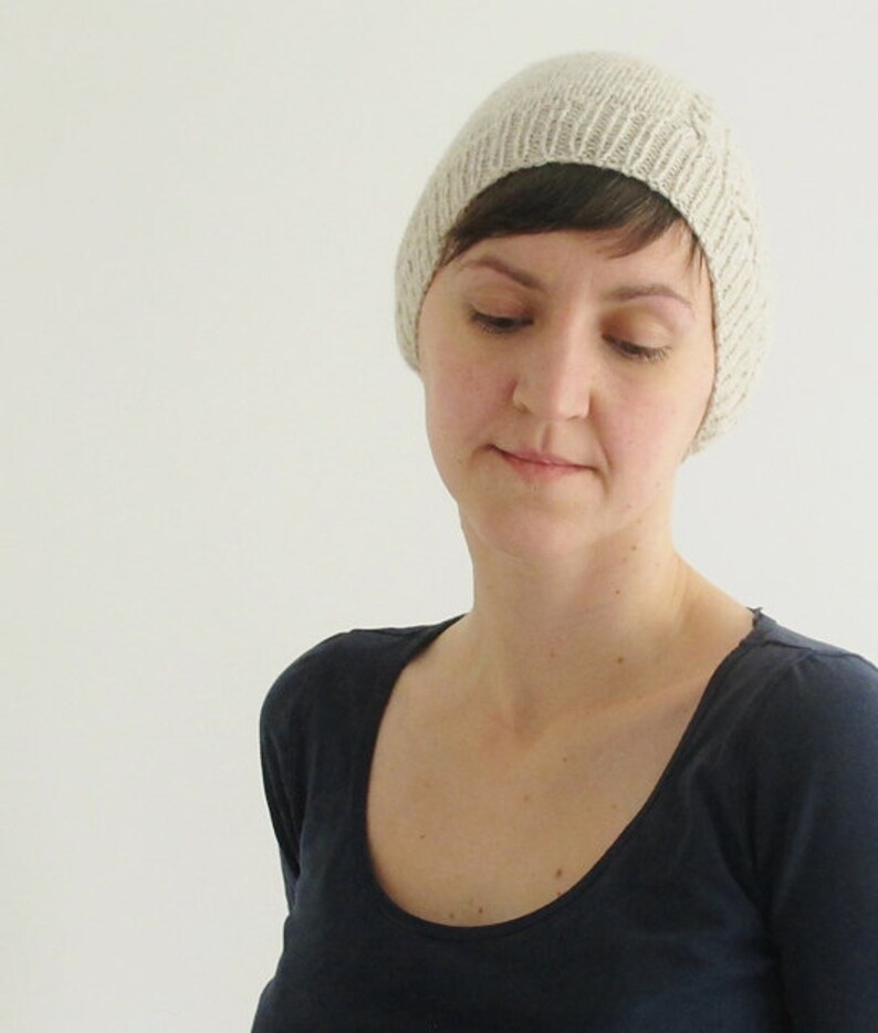 Puede incluir: Una mujer con una boina de punto beige claro con un dise&ntilde;o sencillo. Lleva una camiseta azul oscuro de cuello redondo.