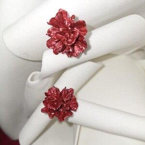 Peut inclure: Deux ronds de serviette rouges en forme de fleur avec une finition pailletée. Les ronds de serviette sont placés sur des serviettes en tissu blanches, créant un contraste de couleurs et de textures. Le design floral ajoute une touche d'élégance.