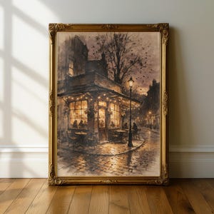 May include: A framed wall art print depicting a cozy corner cafe at dusk with warm glowing string lights, a tall street lamp, and silhouettes of people sitting at outdoor tables on a cobblestone street, all encased in an ornate gold frame.