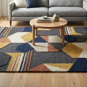 May include: Geometric patterned area rug in shades of gray, navy, yellow, and peach. The rug features a modern design with various geometric shapes and lines. The rug is placed on a hardwood floor in a living room setting.