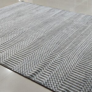 May include: A rectangular area rug with a geometric pattern in shades of gray and white. The rug features a repeating design of diagonal lines and checkered sections, with a fringed edge. The rug is made of a woven material.