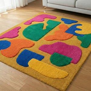 May include: A mustard yellow area rug with abstract shapes in bright colors. The shapes include pink, green, orange, blue, and yellow. The rug is rectangular and sits on a wooden floor.