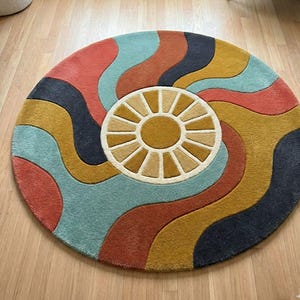 May include: A round rug with a retro design. The rug features a central sunburst pattern in beige and gold, surrounded by wavy stripes in teal, coral, mustard yellow, and navy blue. The rug is made of a soft, plush material.