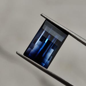 Peut inclure: Une pierre précieuse rectangulaire et facettée, aux teintes bleu foncé, est maintenue par une pince argentée. La taille de la gemme révèle des motifs géométriques internes, créant un jeu d'ombre et de lumière. Un bijou remarquable.