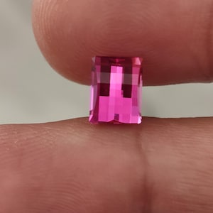 May include: A faceted, rectangular-cut, pink gemstone. The gem is held between two fingers, showcasing its vibrant color and intricate facets. The gemstone's shape is a rectangle with cut corners, reflecting light to create a sparkling effect.