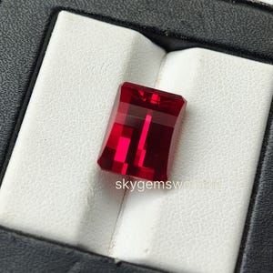 May include: A rectangular, faceted, deep red gemstone with a brilliant lustre and sharp edges. The stone is positioned in a white display slot within a black case. The text "skygemsworldIN" is printed in small white letters on the stone's surface.