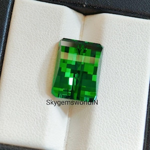 Peut inclure: Une pierre précieuse rectangulaire vert émeraude avec une taille à facettes. La gemme est présentée sur une surface blanche texturée dans une boîte noire. Le texte "SkygemsworldlN" est visible.