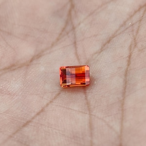 May include: A rectangular, faceted gemstone with a vibrant orange hue. The gem is held in a palm, showcasing its clarity and cut. The gemstone's facets reflect light, creating a sparkling effect. This gemstone is likely used in jewelry.