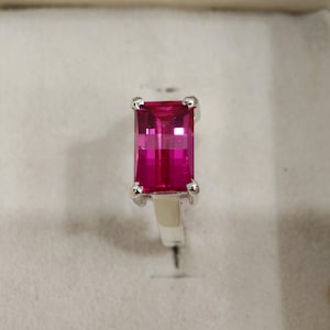 May include: A silver ring featuring a large, rectangular, bright pink gemstone. The gemstone is set with four prongs and has a faceted surface. The ring is displayed in a light-colored jewelry box.