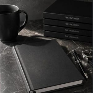 Könnte beinhalten: Ein schwarzes Notizbuch mit einem Lesezeichen liegt offen auf einer Marmoroberfläche, neben einem Stift und einer Uhr. Ein Stapel Notizbücher mit der Aufschrift "THE UNCOMMON" und eine schwarze Tasse sind ebenfalls zu sehen. Das Bild hat eine monochrome Ästhetik.