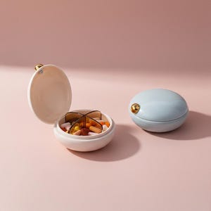 Puede incluir: Dos pastilleros, uno abierto y otro cerrado, sobre una superficie rosa. El organizador abierto es blanco con un interior dividido que contiene varias pastillas. El organizador cerrado es azul claro con un cierre dorado.