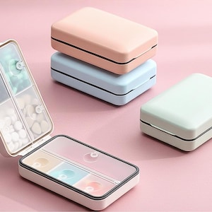 Può includere: Una collezione di portapillole dai colori pastello. Uno è aperto, rivelando scomparti pieni di pillole. Gli organizzatori chiusi sono in tonalità di rosa, blu e verde, con finiture nere. Lo sfondo è rosa tenue.