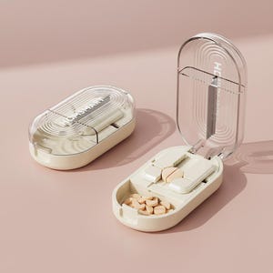 Puede incluir: Dos pastilleros beige con tapas transparentes. Uno está abierto, revelando pastillas y un cortador de pastillas. El otro está cerrado. El texto "HIGH SMART" es visible en la tapa del pastillero cerrado.