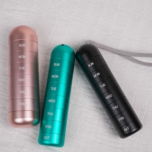 Puede incluir: Tres pastilleros cilíndricos en oro rosa, verde azulado y negro. Cada pastillero está dividido en siete secciones etiquetadas para cada día de la semana, con los días de la semana impresos en blanco. Un cordón gris está unido al pastillero negro.